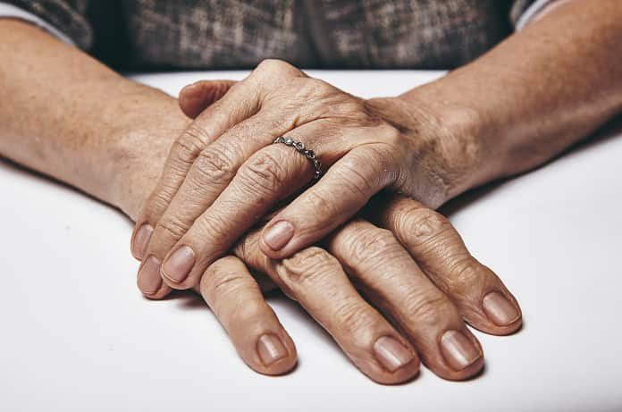 macro-of-an-old-lady-sitting-with-her-hands-clasped-on-a-table-elderly-womans-hands-with-a-ring-resting-on-grey-surface-focus-on-hands_HpbS7v84tx-min-696×461
