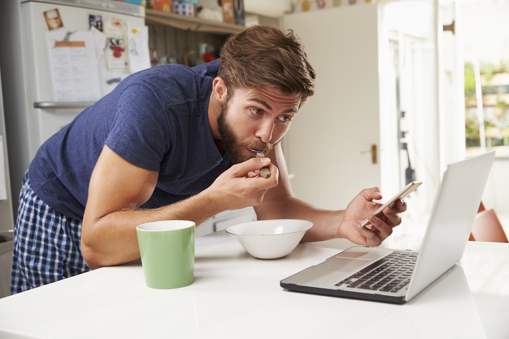 Man Eating Breakfast Whilst Using Mobile Phone And Laptop